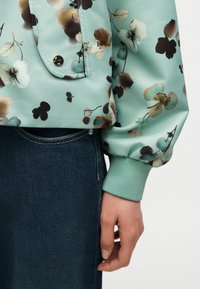 Main reposant sur le côté portant un haut à manches longues floral vert clair avec poche pression et une jupe en jean bleu foncé.