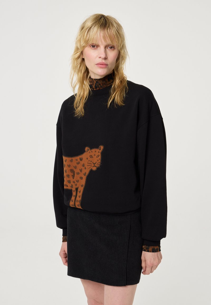 Blonde Frau, die einen schwarzen Pullover mit großem braunem Leopardenmotiv, einen dunkel gemusterten Rollkragenpullover und einen schwarzen Cordrock vor einem weißen Hintergrund trägt.