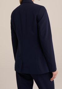 Navy blejzr na míru se strukturovanými rameny, jedním rozventilováním a hladkou texturou. Obsahuje klopový límec a přiléhavý střih.