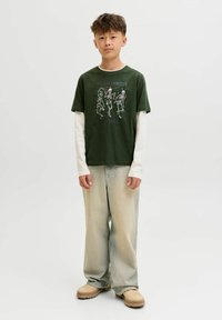 Adolescent portant un T-shirt vert foncé avec des squelettes et du texte, des manches longues blanches en dessous, un jean clair ample et des chaussures beiges, debout.