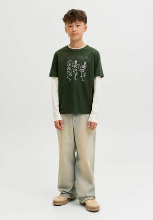 Tiener in een donkergroene T-shirt met skeletten en tekst, daaronder een witte lange mouw, losse lichtgewassen spijkerbroek en beige schoenen, staand.