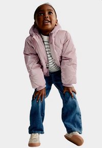 Lichtroze gewatteerd jack met diagonaal geruit patroon, gestreept wit-zwart shirt, blauwe spijkerbroek en beige sneakers met rubberen zolen.