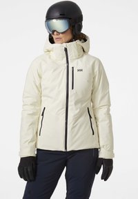 Witte skijack met een capuchon, voorzien van zwarte ritsen en zakken. Gecombineerd met zwarte handschoenen en een zwarte helm met goggles.