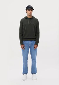 Mørkegrønn hettegenser med kengurulomme, kombinert med lyseblå jeans og hvite joggesko. En svart caps fullfører antrekket.