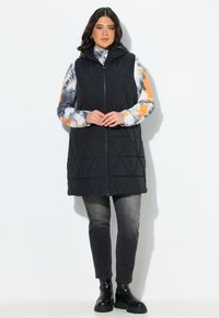 Svart vadderad väst med huva, med en framzipper, bärs över en flerfärgad tie-dye långärmad skjorta och matchas med mörka jeans och stövlar.