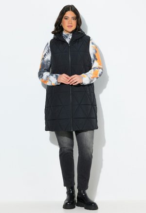 Gilet trapuntato nero con cappuccio, caratterizzato da una zip frontale, indossato sopra una camicia a maniche lunghe tie-dye multicolore e abbinato a jeans scuri e stivali.