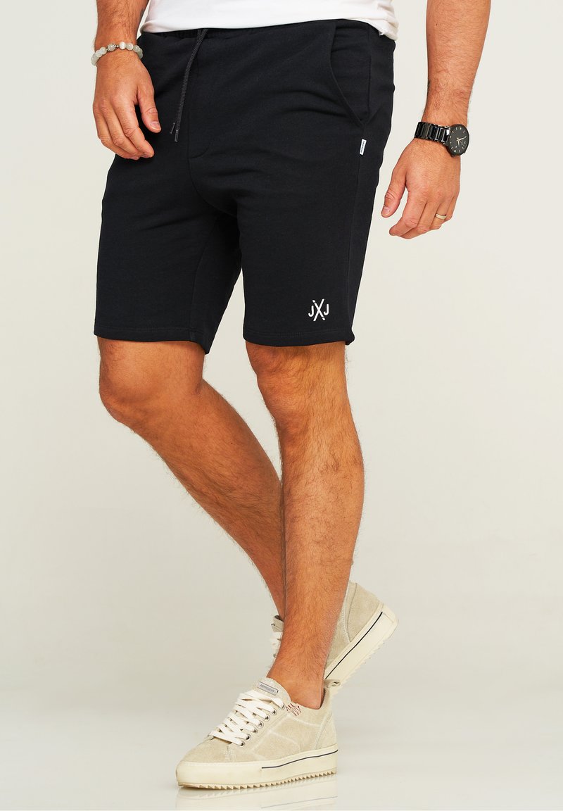 Black cotton shorts with a drawstring waist, side pockets, and a small white logo. Paired with light-colored, textured sneakers.
