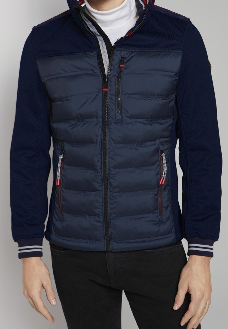 Homme portant une veste matelassée bleu marine avec poches à fermeture éclair sur la poitrine et les côtés, associée à un pantalon noir et un col roulé blanc.