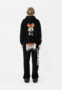 Sudadera negra con gráfico de oso de dibujos animados en la parte posterior, combinada con pantalones de chándal negros que presentan texto blanco y un diseño de oso; ambos en un corte relajado.