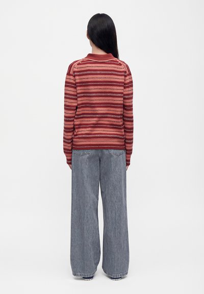 Personne aux longs cheveux foncés vue de dos, portant un pull rayé rouge et un jean gris à jambes larges sur un fond blanc uni.