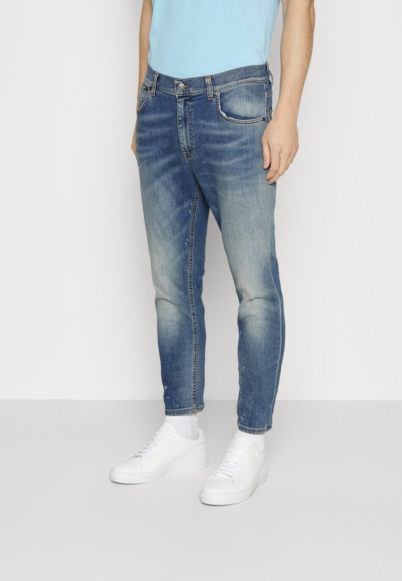 Homme portant un jean bleu délavé, des chaussettes blanches et des baskets blanches, debout devant un fond clair uni.