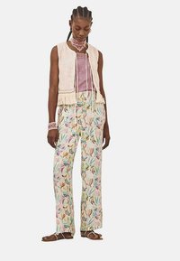 Chaleco de ganchillo sobre una blusa lavanda, combinado con pantalones florales ligeros que presentan hojas y pájaros coloridos. Los acentos incluyen una cinta y sandalias.