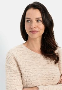 Helles Beige, strukturiertes Strickpullover mit weitem Ausschnitt und gerippten Akzenten, getragen von einem Model mit dunklem, welligem Haar und natürlichem Make-up.