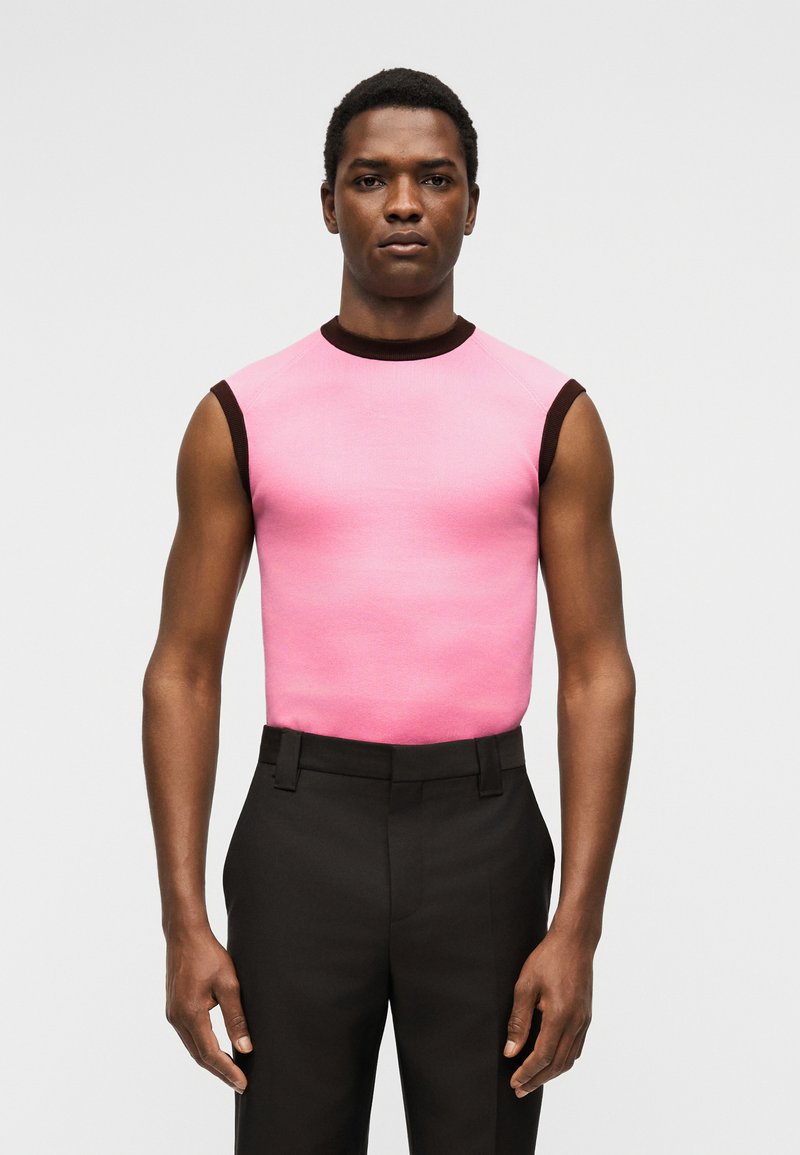 Man standing against plain background wearing sleeveless pink top with dark brown trim and high-waisted black trousers.