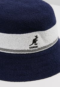 Chapeau seau bleu marine en tissu éponge, avec une bande blanche et un accent gris, portant un logo de kangourou noir sur le côté.