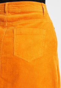 Jupe en velours côtelé orange avec une texture côtelée verticale, une poche arrière et une taille ajustée. Le tissu semble doux et durable.