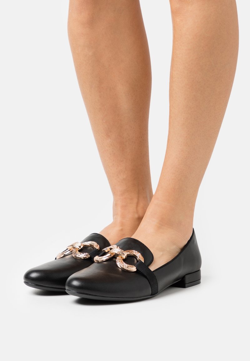 Chaussures plates en cuir noir avec un talon plat, dotées de deux accents en chaîne dorée à l'avant et d'une texture lisse.