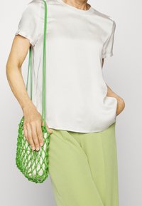 Haut beige clair à manches courtes, associé à un pantalon large vert pâle. Tient un petit sac tressé vert au design texturé.