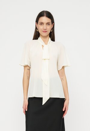 Γυναίκα με μακριά σκουρόχρωμα μαλλιά που φοράει λευκό μπλουζάκι με κοντά μανίκια, με δέσιμο μπροστά και πιετέ λεπτομέρειες, σε συνδυασμό με μαύρη φούστα.