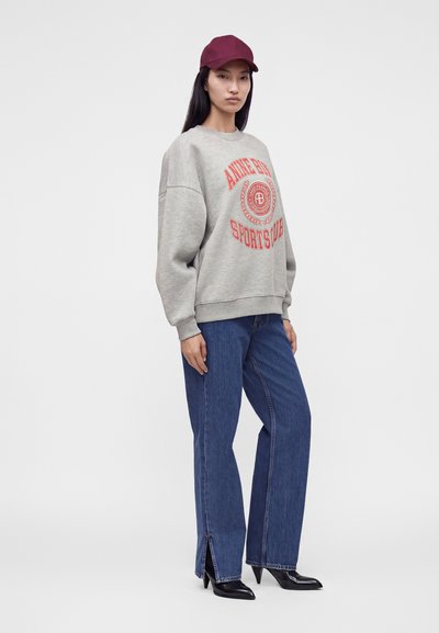 Femme portant un sweat-shirt gris oversize avec texte rouge, jean bleu à jambes larges avec fentes aux chevilles, talons noirs et casquette de baseball bordeaux.