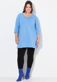 Himmelblaues Tunika mit V-Ausschnitt und Dreiviertelärmeln, kombiniert mit schwarzen Leggings und blauen Reißverschluss-Stiefeletten mit Absatz. Glatte Stoffstruktur.