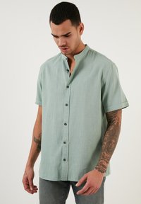 Camisa de manga corta, con cuello, en tela verde claro, que presenta un cierre de botones y un corte casual. La textura es suave con un sutil patrón de tejido.