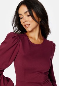 Bordeaux top met pofmouwen, gladde textuur, ronde halslijn en een aansluitend ontwerp dat de schouder- en armgebieden accentueert.