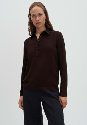 Donna con capelli castano chiaro che indossa una polo marrone scuro a maniche lunghe e pantaloni neri, in piedi davanti a uno sfondo chiaro uniforme.