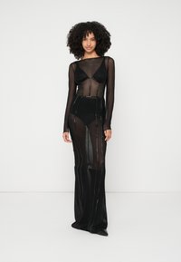 Juoda permatoma suknelė ilgomis rankovėmis, su subtiliomis vertikaliomis linijomis, prigludusiu liemenėliu ir grindų ilgio sijonu su skaidria medžiaga.