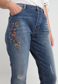 Siniset farkut, joissa sivutasku, kukkakirjailu ja kulutuspintaiset yksityiskohdat. Keskikorkea vyötärö, nappikiinnitys ja vyölenkit.