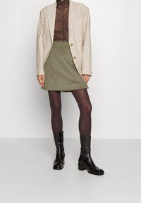 Beige blazer, doorzichtig bruin top met gerimpelde textuur, olijfgroene rok, donkerbruine pantyhose en zwarte glanzende enkellaarsjes met een blokhak.