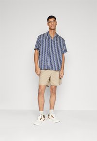 Hombre con una camisa de manga corta azul con estampado, pantalones cortos beige, calcetines blancos y zapatillas blancas, de pie contra un fondo sencillo.