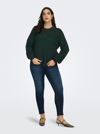 Femeie cu păr lung și închis la culoare, purtând un pulover verde închis tricotat, blugi skinny albastru închis și pantofi verzi măsliniu cu toc mic, stând pe un fundal simplu.