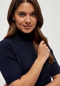 Mlada žena s dugom smeđom kosom koja nosi tamnoplavi pulover kratkih rukava s visokim ovratnikom, držeći pramen kose na jednostavnoj pozadini.