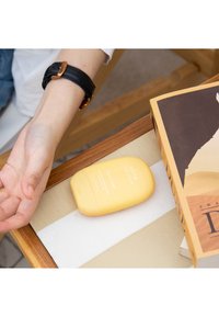 Contenedor de crema de manos amarilla etiquetado como "Coco Cooler" sobre una superficie texturizada, junto a un libro abierto, con una mano extendiéndose hacia él.