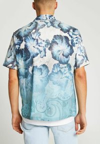 Camisa de manga corta con un patrón floral azul, fondo azul claro y sutiles detalles de paisley. Fabricada con un tejido ligero.