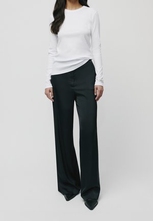 Femme portant une chemise blanche à manches longues, un pantalon large noir et des chaussures noires pointues, debout devant un fond gris uni.