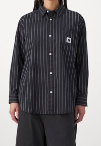 Camisa negra de botones con rayas verticales blancas, mangas largas y un bolsillo en el pecho con un logo. Tela ligera con una textura suave.