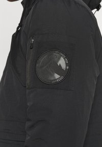 Veste isolante noire avec une poche zippée sur la manche. Présente un écusson rond et texturé représentant des montagnes et des éléments de ski.