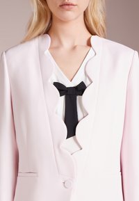 Vaaleanpunainen bleiseri, jossa on aaltoilevat reunat, yksittäinen napitus ja taskuyksityiskohdat, valkoisen paidan päällä, jossa on musta solkiyksityiskohta.