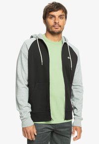 Čierna a šedá zipsová mikina s kapucňou, dvoma prednými vreckami a svetlozeleným tričkom pod ňou. Mäkká tkanina s voľným strihom.