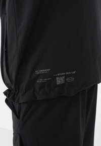 Giacca in tessuto tecnico nero con una texture liscia, caratterizzata da un orlo arricciato, etichette stampate e un codice QR sul lato inferiore.