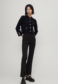 Chaqueta negra texturizada con cierre de botones, combinada con pantalones negros de cintura alta que presentan aberturas en el dobladillo. La modelo lleva zapatos negros.