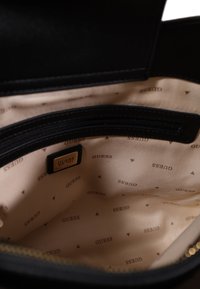 Interno della borsa nera con tessuto liscio, caratterizzato da una fodera beige con logo "GUESS" ripetuto e piccoli accenti a triangolo. Inclusa tasca con zip.