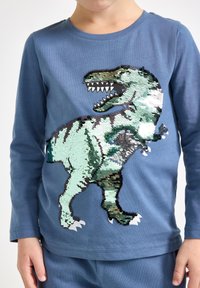 Langärmliges blaues Hemd mit einem T-Rex-Design aus umkehrbaren Pailletten, mit grünen und schwarzen Details sowie strukturiertem Stoff.