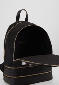 Black backpack with gold zippers, suede-like top, textured bottom, and a spacious interior, featuring a label and a flat pocket inside.