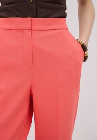Coral trousers with a smooth texture, featuring a tailored fit, side pockets, and a flat front. Includes a brown top and wrist accessory.