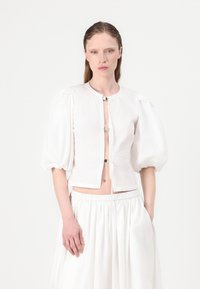 Blusa bianca cropped con maniche a sbuffo, chiusura anteriore con bottoni e un tessuto leggero. Caratterizzata da un collo alto e un design minimalista.