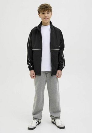 Veste zippée noire avec des garnitures blanches, associée à un t-shirt blanc, un jean gris et des baskets noir et blanc. Le modèle se tient devant un fond uni.