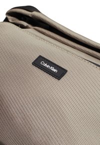 Beige szövet táska texturált felülettel, amelyen fekete Calvin Klein logó folt látható. A dizájnon cipzár és lekerekített szélek találhatók.